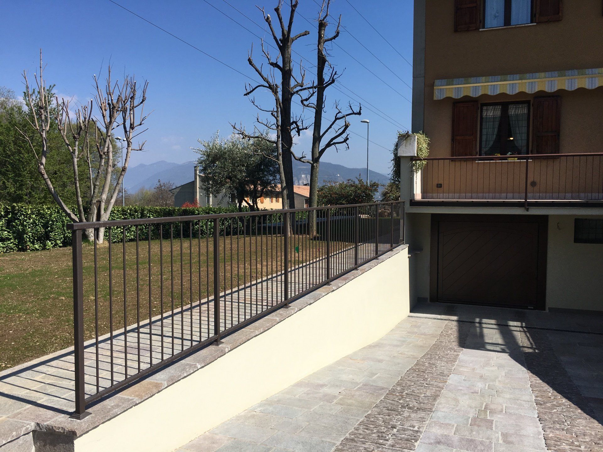 Passeggiata esterna soleggiata accanto a un edificio con ringhiera in metallo nero e alberi sullo sfondo.