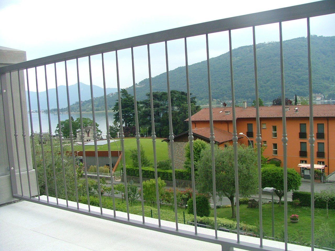 Ringhiera del balcone con vista su verdi colline, alberi e un edificio arancione sotto un cielo nuvoloso.