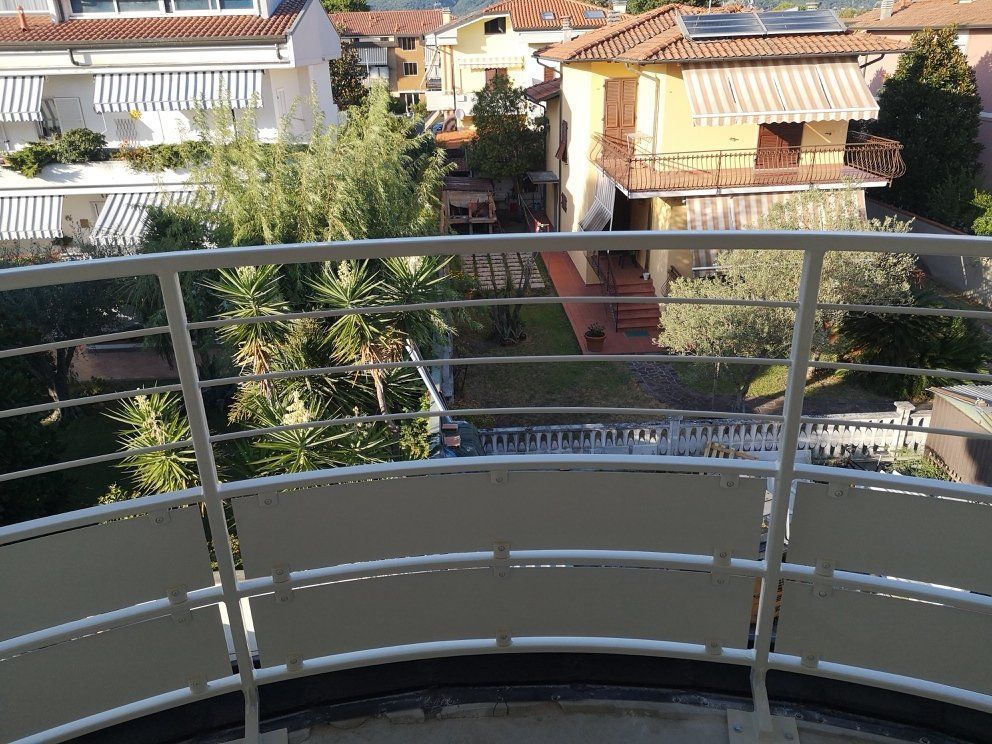 Vista da un balcone che si affaccia su case di periferia, alberi e una stradina stretta, in pieno giorno.