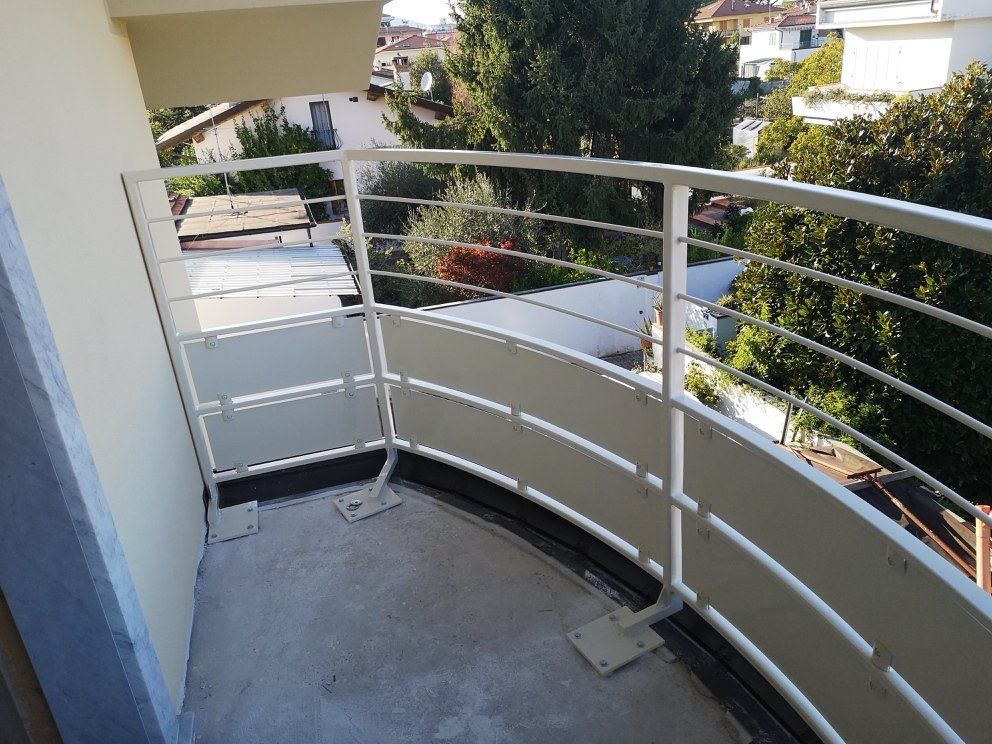 Balcone curvo con ringhiera bianca e pavimento in cemento affacciato su una strada residenziale e sul verde.