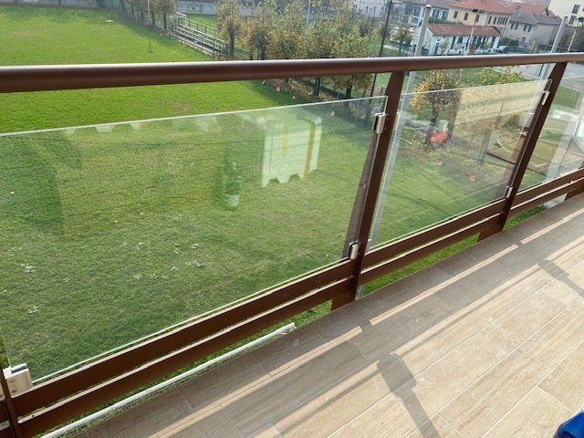 Balcone con ringhiera in vetro affacciato su un prato verde e su case in lontananza in una giornata di sole.