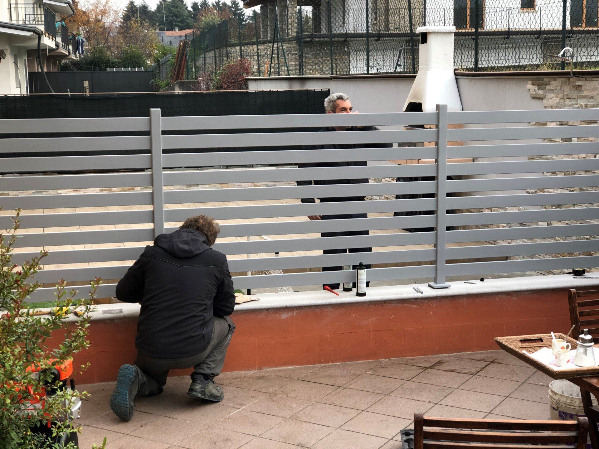 Una persona è inginocchiata accanto a una recinzione a doghe grigie su un patio piastrellato, con un cane bianco che sbircia da sopra.