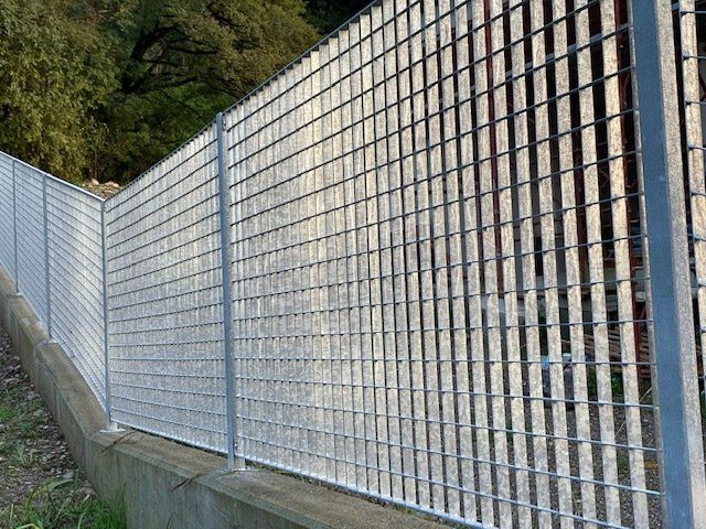 Recinzione in rete metallica lungo un muro di cemento inclinato accanto agli alberi