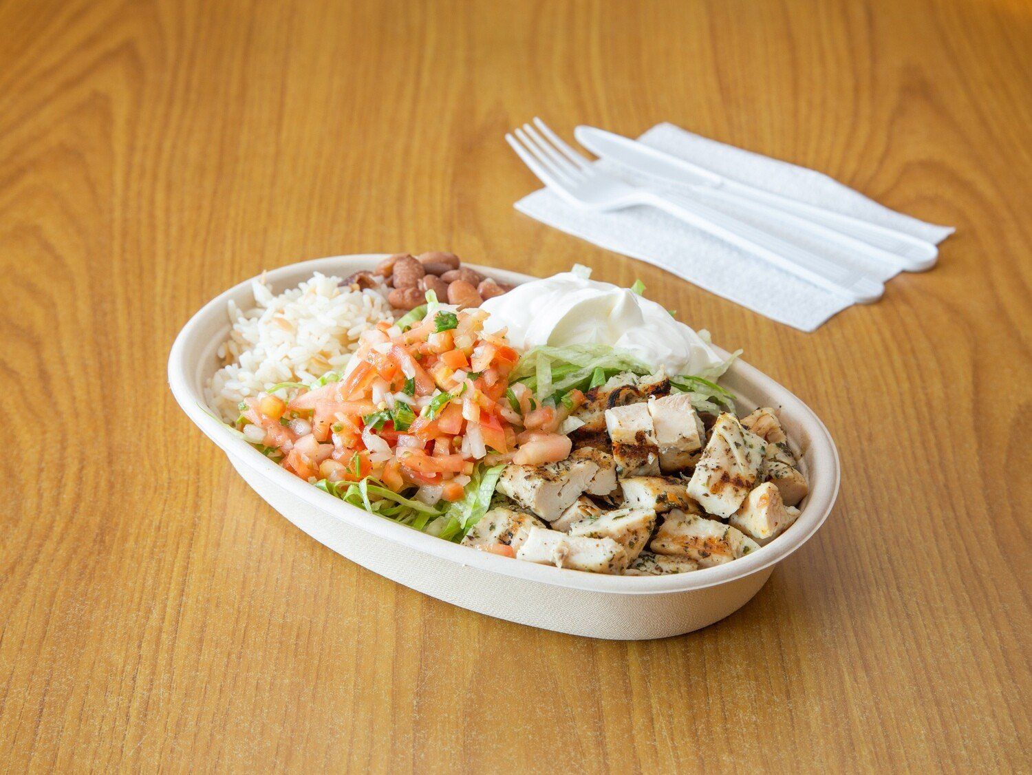 A bowl of food is on a wooden table next to a napkin and fork.