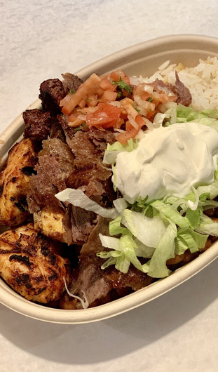 A bowl of food with meat , chicken , lettuce and sauce on a table.