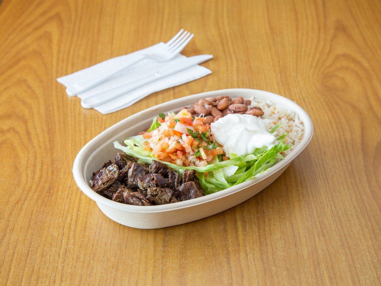 A bowl of food is sitting on a wooden table next to a napkin and fork.