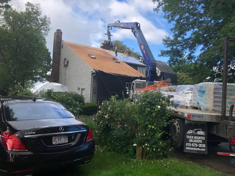 Blue crane lifting roofing materials onto a house with a damaged roof; car and truck in foreground.