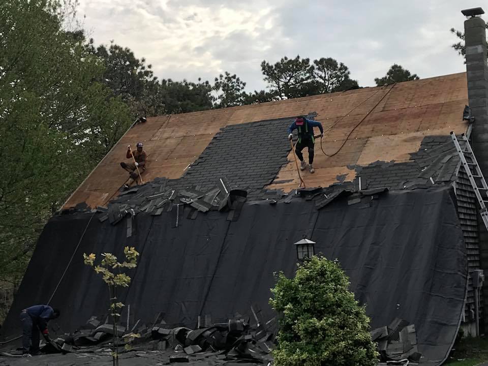 Workers on a roof installing a dark tarp over storm-damaged shingles under cloudy skies
