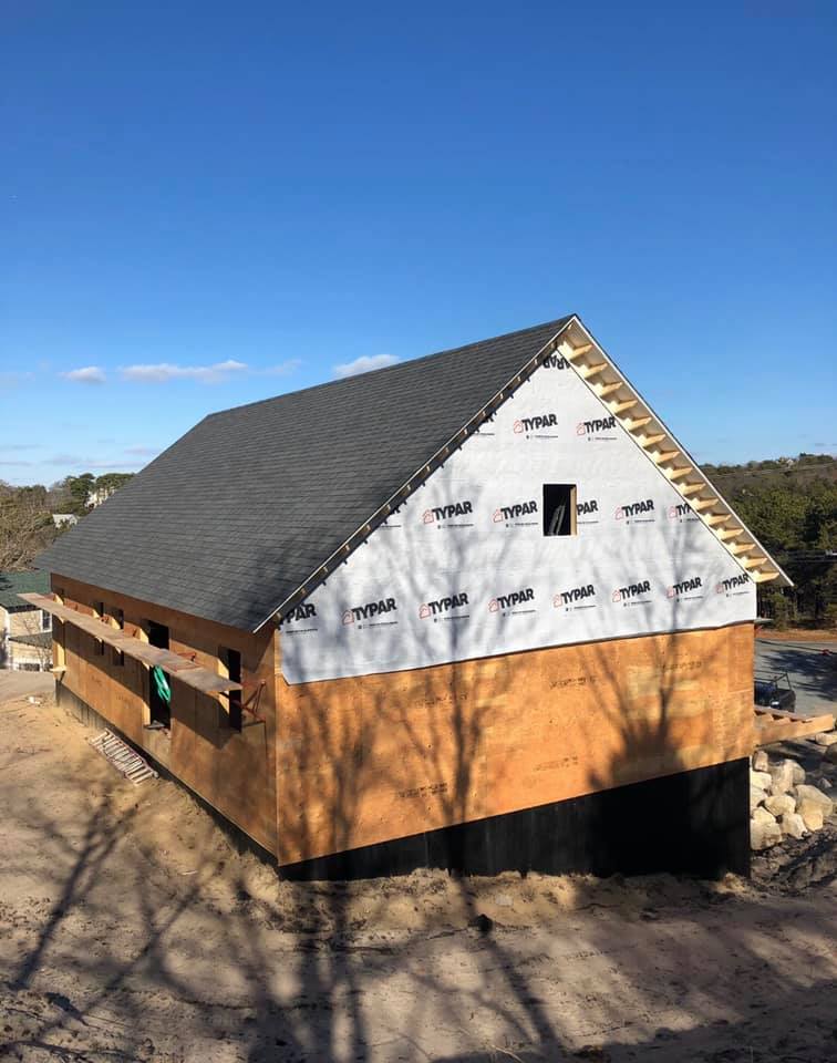 House under construction with gray roof, white wrap, and brown siding in a sunny wooded area