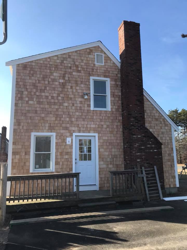 Small shingled house with white trim, brick chimney, and front door in daylight.