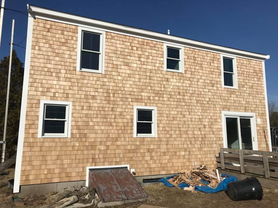 Two-story tan shingled house exterior with white-trimmed windows and construction debris in front