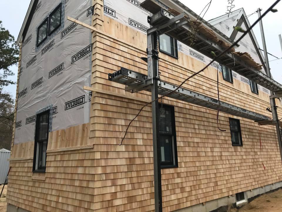 Partially clad house under construction with wood shingles, wrapped upper walls, and scaffolding on the side