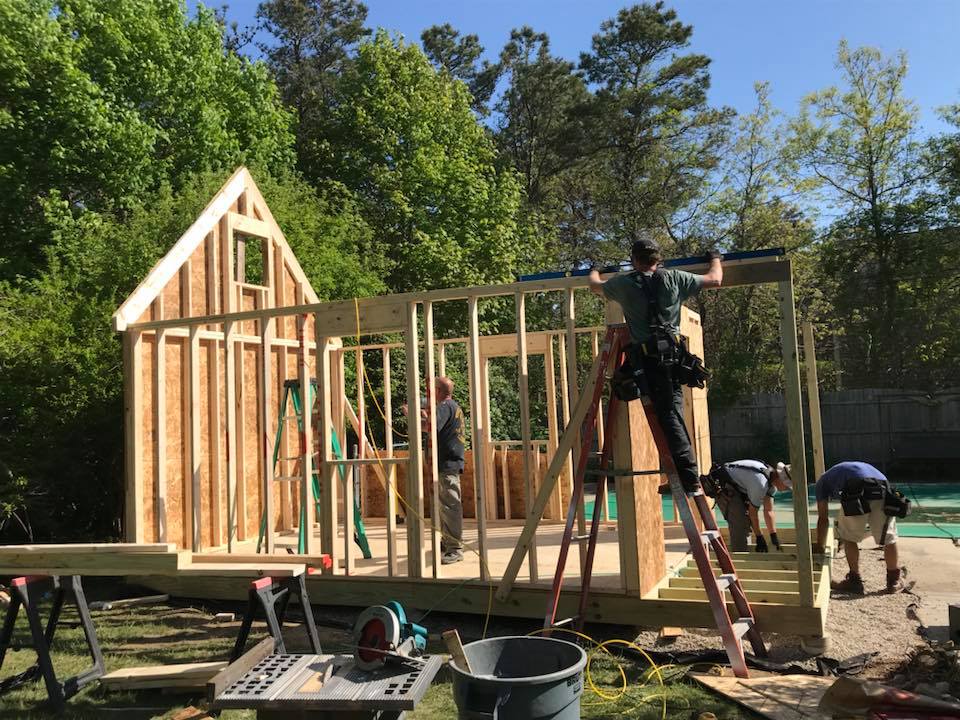 Builders framing a small wooden house outdoors, with ladders, tools, and trees in the background