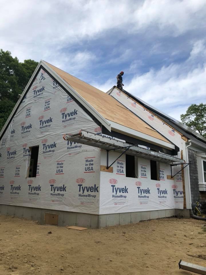 House under construction wrapped in Tyvek, with a worker on the roof under a cloudy sky