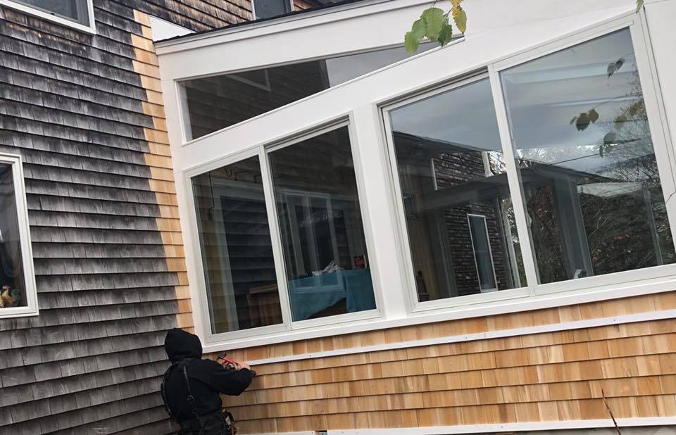 Person in a black hoodie standing by a shingled house with large windows and a brick foundation.