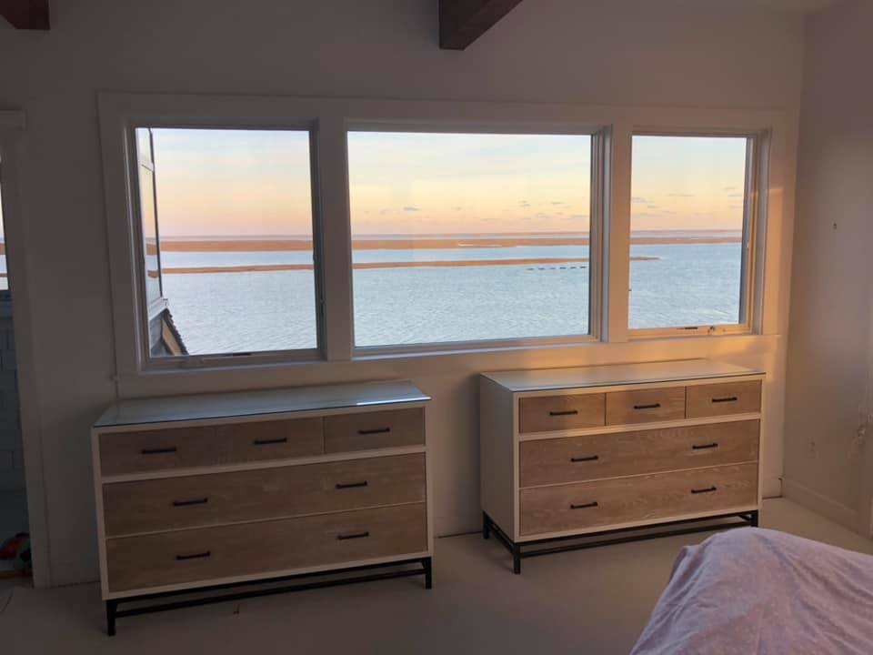 Bedroom with two dressers beneath a large window overlooking calm water at sunset