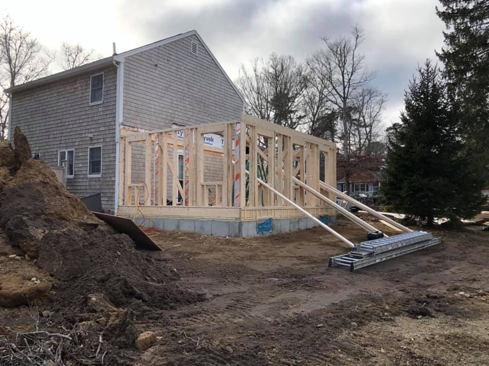 House under construction with exposed wooden framing and roof, concrete foundation, and dirt yard outside