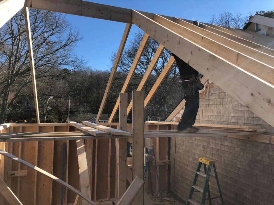 Worker framing a roof on a wooden house under construction, with a ladder and blue sky.
