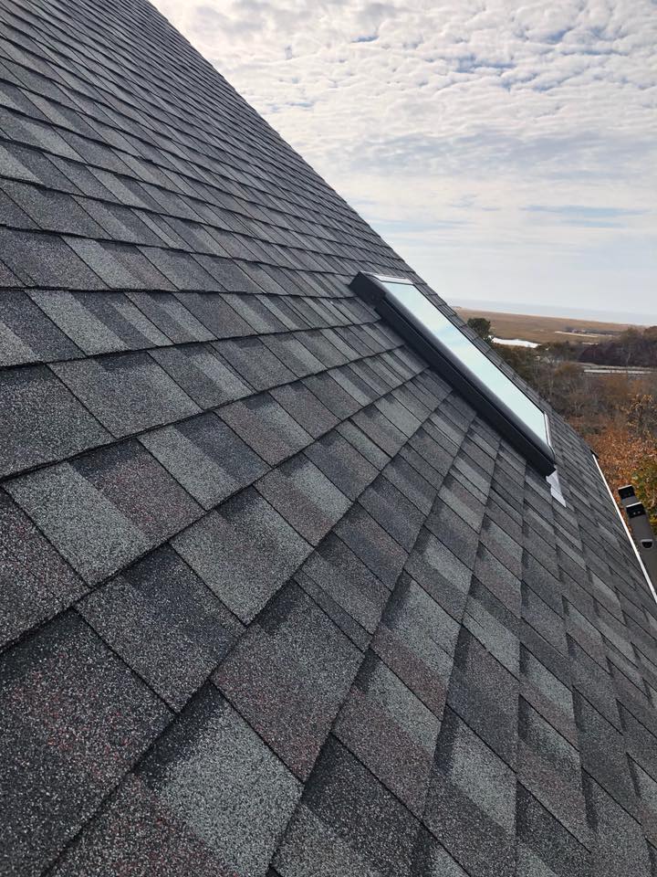 Steep gray shingled roof with a skylight, viewed against a cloudy sky and distant landscape