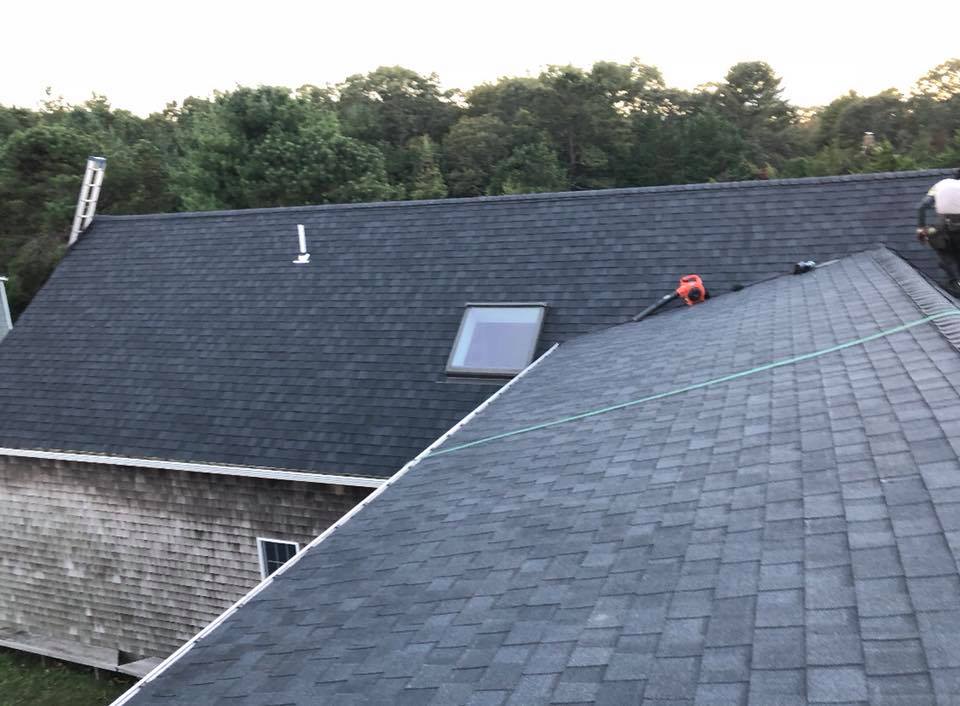Two dark-gray shingle roofs, one with a skylight, with trees in the background and a red object on the roof ridge