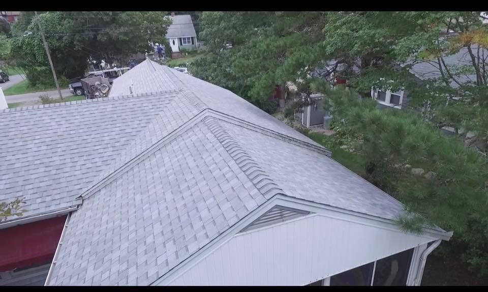 Aerial view of a gray shingled house roof surrounded by green trees and neighboring homes.