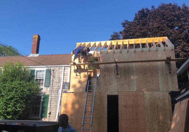 Roof workers building an extension beside a house, with ladder and exposed wooden framing.
