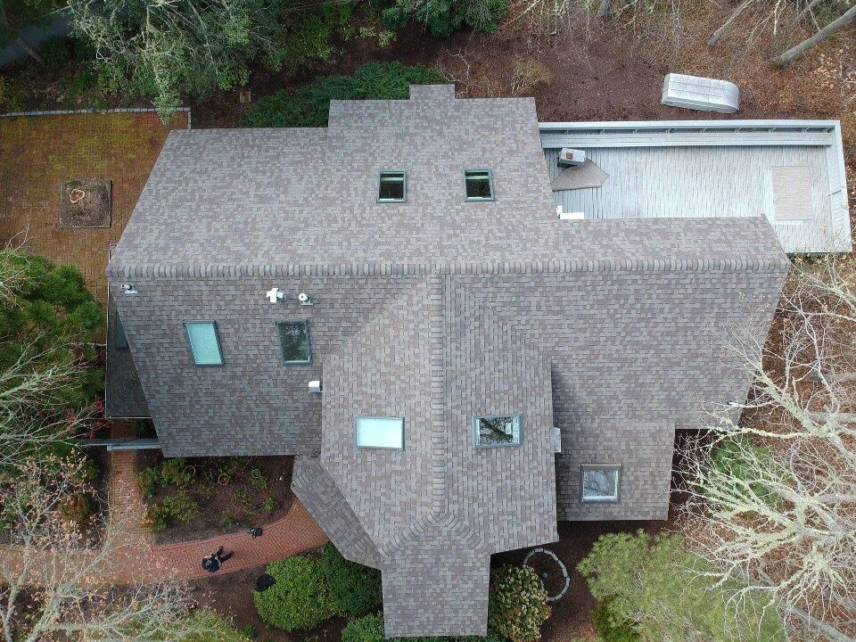 Aerial view of a gray-shingled house with multiple roof sections and surrounding trees