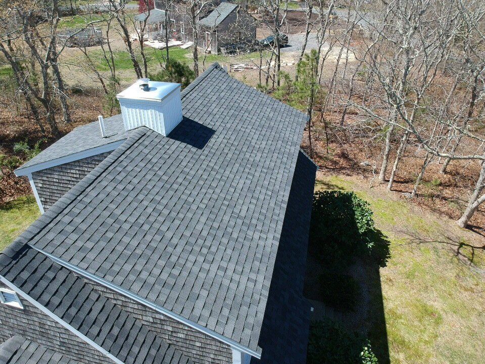 Aerial view of a house roof with dark shingles and a small white chimney, surrounded by trees and grass.