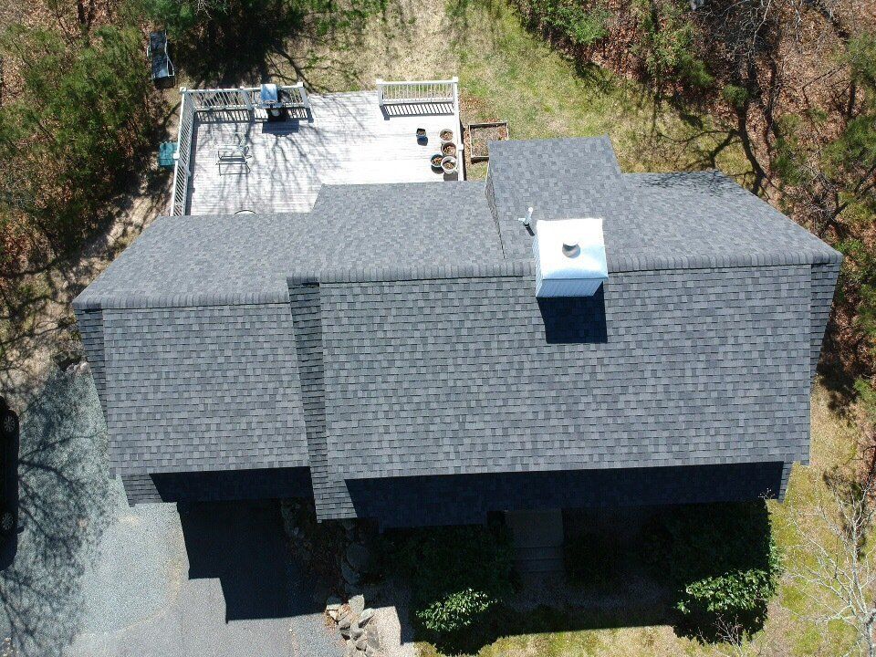 Aerial view of a gray-shingled house with a flat white rooftop deck and skylights, surrounded by trees.