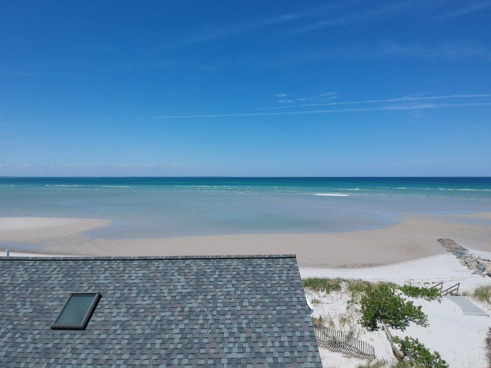 Sandy beach with calm blue ocean and sky, seen past a gray shingle roof in the foreground
