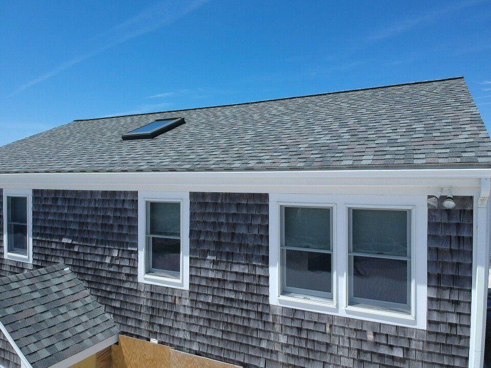 Gray shingled house roof with skylight and white-trimmed windows under a blue sky