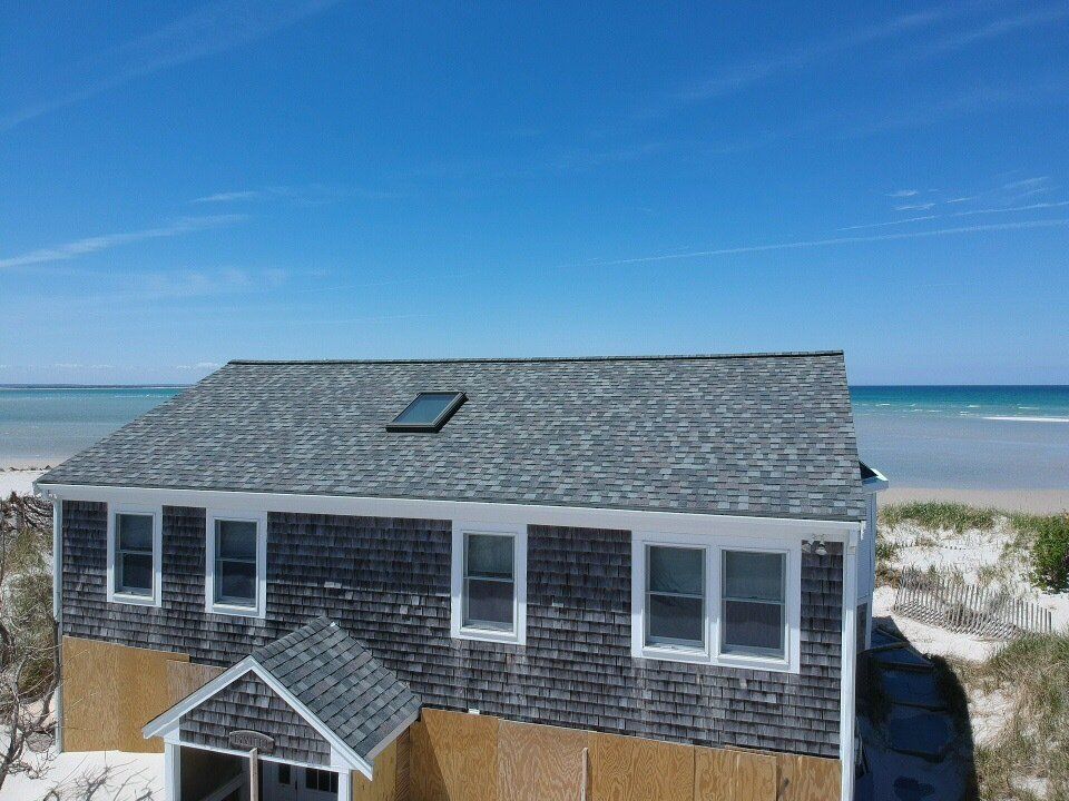 Beachfront house with gray shingle roof and white trim, overlooking sand dunes and blue ocean under clear sky