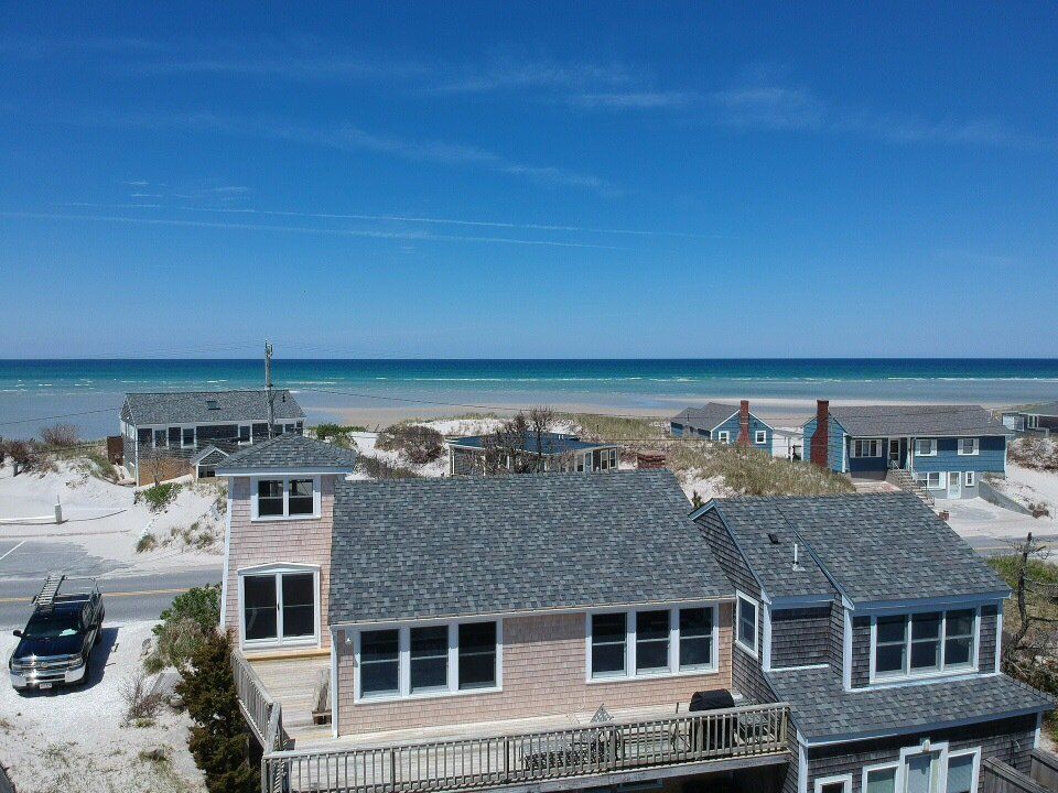 Beachfront house with gray roof and white siding overlooking sand dunes and ocean under a clear blue sky