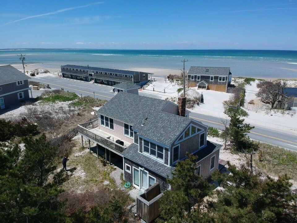 Beachfront house with gray roof and white trim, overlooking dunes, ocean, and nearby homes.