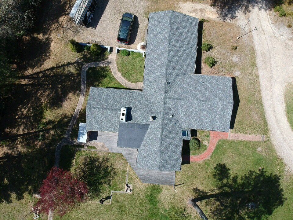 Aerial view of a gray-roofed house with driveway, cars, and trees in a suburban yard