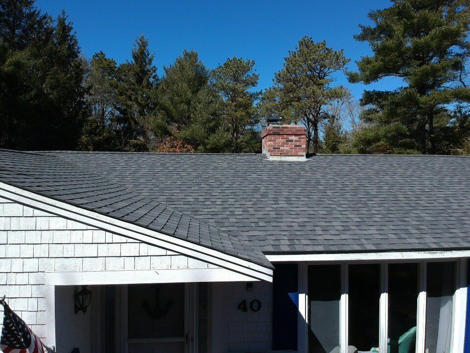 Suburban house roof with gray shingles, brick chimney, white siding, and trees against a blue sky