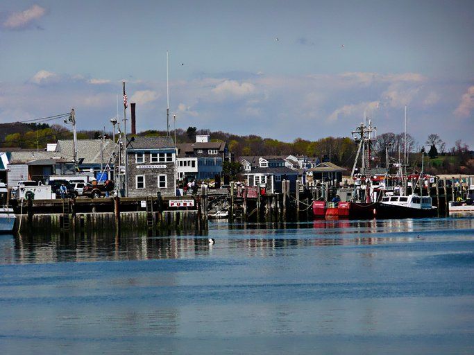 Harbor with docked fishing boats, waterfront buildings, and calm blue water under a cloudy sky