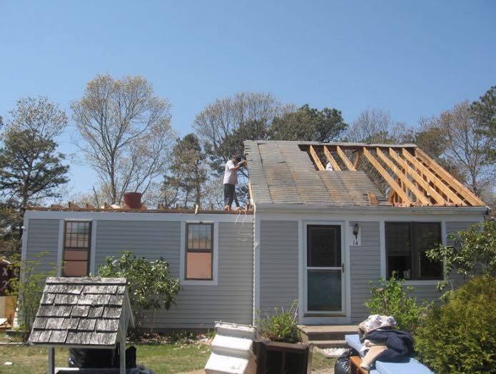 Workers reroofing a gray house, with exposed rafters and shingles removed on the right side.