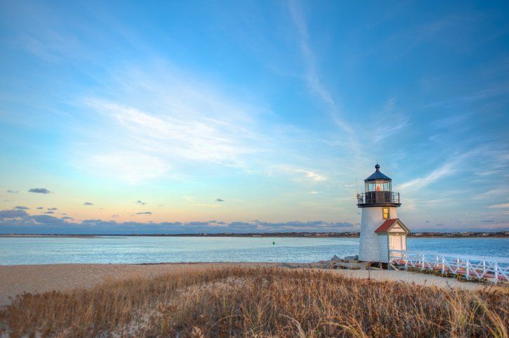 Lighthouse on a grassy shoreline at sunrise with calm water and a colorful sky