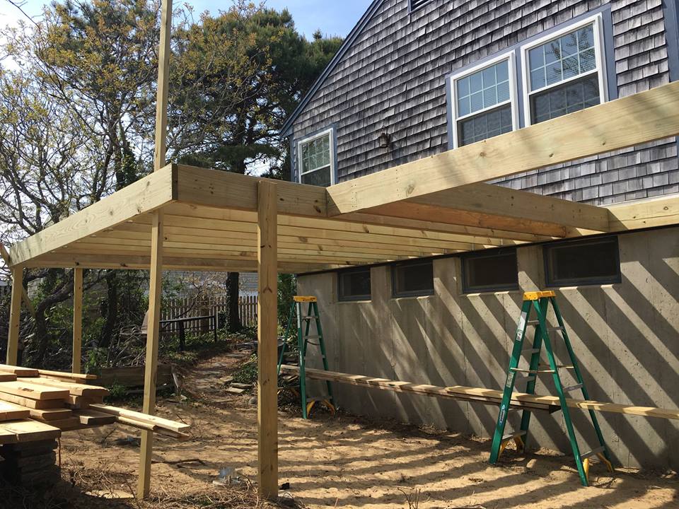 Wooden deck under construction beside a house, with posts, beams, ladder, and stacked lumber.
