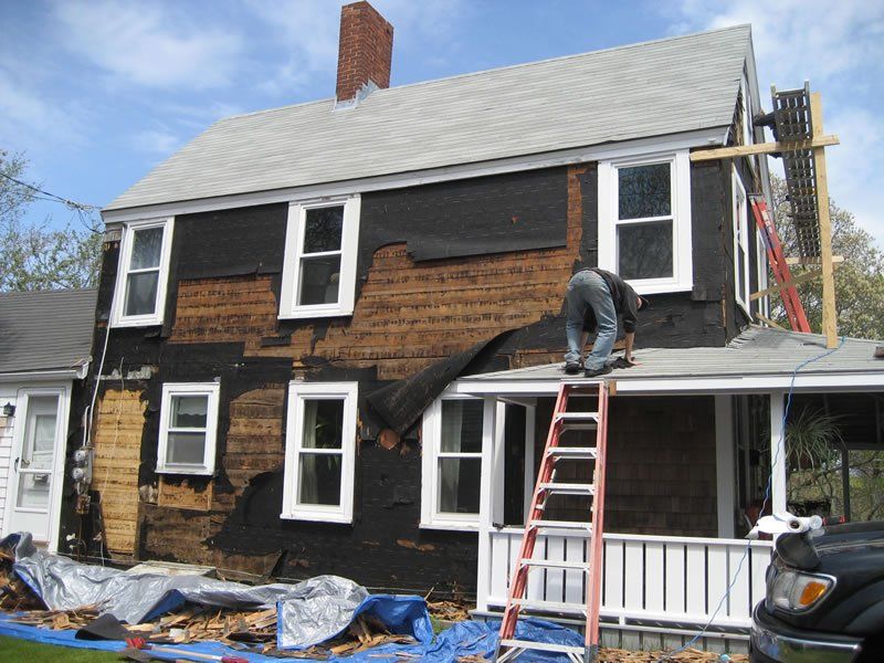 Worker on ladder repairing the roof and siding of a black two-story house with white trim.