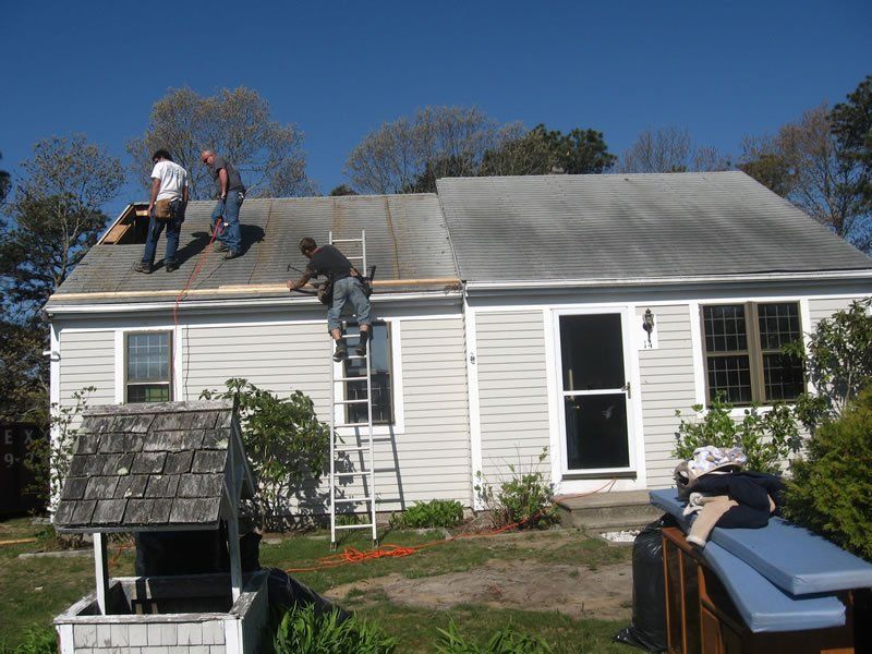 Workers repairing a white house roof, with ladder and tools in the yard