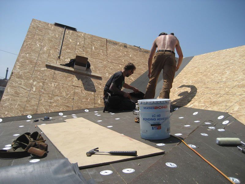Two people working on a sloped rooftop with tools and a white bucket under a blue sky