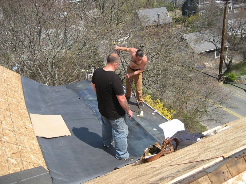 Two workers installing roofing underlayment on a sloped roof, with plywood and tools nearby.