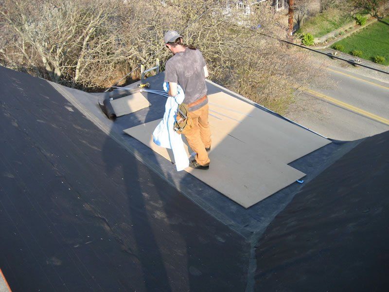 Worker installing roofing material on a flat roof near tree-lined street