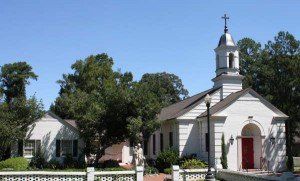 White church building with steeple, red door, and surrounding trees under a blue sky.