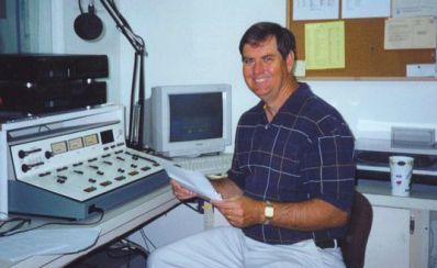 Man seated at a radio control panel, holding papers. He smiles in a radio studio with a computer and microphone.