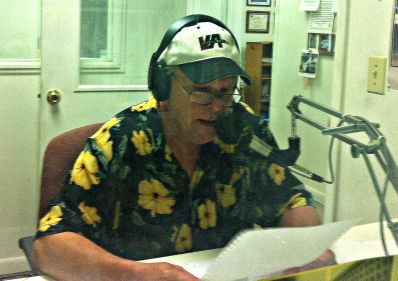 Man in floral shirt and hat speaking into microphone at a radio studio, reading from papers.