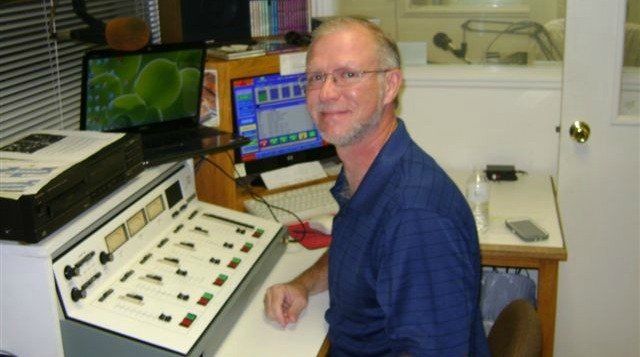 Man sits at a desk, smiling. He’s in a control room with audio equipment and computers.