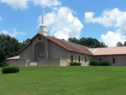 Tan brick church with white steeple on green lawn under blue sky.